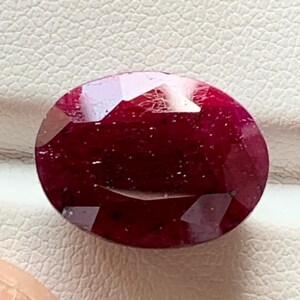 Puede incluir: Un rubí facetado de forma ovalada. La gema es de un color rojo intenso, con inclusiones internas visibles. El rubí se exhibe sobre una superficie blanca, mostrando su brillo y claridad. La gema es sostenida por una mano.