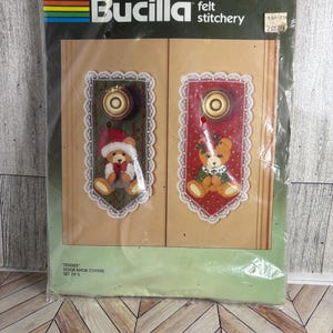 May include: A Bucilla felt stitchery kit for door knob covers. The kit includes two felt covers with teddy bear designs, one in green and one in red, with lace trim. The package has the text "TEDDIES" and "DOOR KNOB COVERS SET OF 2".