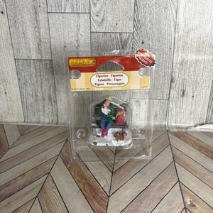 May include: A Lemax figurine in a clear plastic package. The figurine depicts a person sitting on a bench with a child and a squirrel. The person is wearing a green jacket and blue pants. The package has the text "Figurine" in multiple languages.
