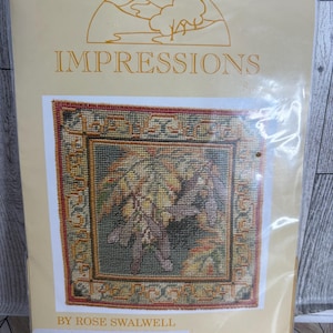 May include: A craft kit titled "Impressions" by Rose Swalwell. The kit features a needlepoint design with a central image of autumn leaves in shades of brown, green, and yellow, framed by a decorative border. The kit is sealed in a clear plastic bag.