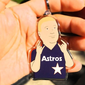 May include: A clear acrylic keychain featuring a cartoon character wearing a purple jersey with the word "Astros" and a white star.