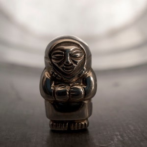 May include: A silver figurine of a person in a hooded garment, hands clasped. The figure has a smiling face and is likely a small statue or ornament. The background is blurred, with a dark surface below.
