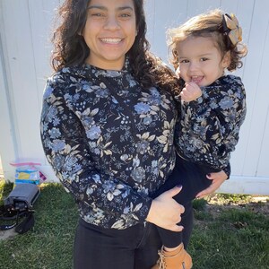Peut inclure: Une femme et une jeune fille portant des sweats à capuche à imprimé floral assorti. La femme tient la fille dans ses bras. La fille porte des bottes brunes.