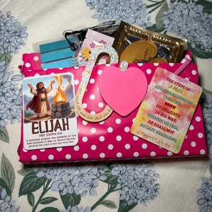 May include: A pink gift box with white polka dots featuring a sticker of Elijah, a floral-patterned hook, a solid pink heart-shaped card, and a rectangular card with the text The steps of a man are established by the Lord, Psalm 37:23-24.