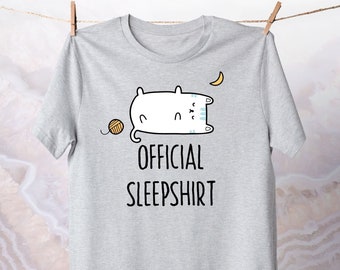 May include: Light gray t-shirt with the text "OFFICIAL SLEEPSHIRT" in black. The shirt features a cartoon cat sleeping on a pillow, a crescent moon, and a ball of yarn. The shirt is hanging from a wooden clothespin.