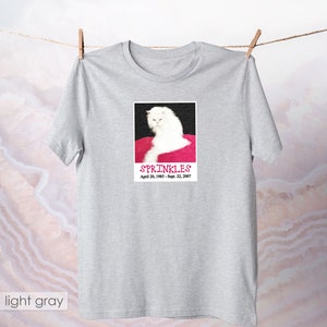 May include: A light gray t-shirt with a white cat on a pink background. The cat is looking at the camera with a serious expression. The text "SPRINKLES" is printed below the cat, along with the dates "April 20, 1985 - Sept. 22, 2007".