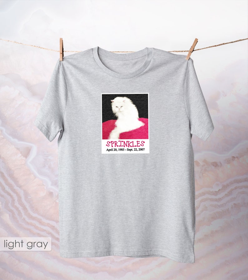 May include: A light gray t-shirt with a white cat on a pink background. The cat is looking at the camera with a serious expression. The text "SPRINKLES" is printed below the cat, along with the dates "April 20, 1985 - Sept. 22, 2007".