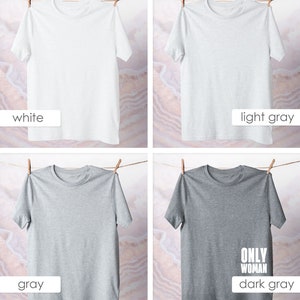 May include: Four t-shirts hanging on a clothesline. The t-shirts are in four different colors: white, light gray, gray, and dark gray. The dark gray t-shirt has the text "ONLY WOMAN" printed on it in white.