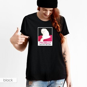 May include: Black t-shirt with a white cat on a pink cushion. The cat is looking to the left. The text "SPRINKLES April 20, 1985 - Sept. 22, 2007" is printed below the cat.