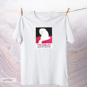 May include: White t-shirt with a photo of a white cat on a pink background. The photo has the text "SPRINKLES" and "April 20, 1985 - Sept. 22, 2007".