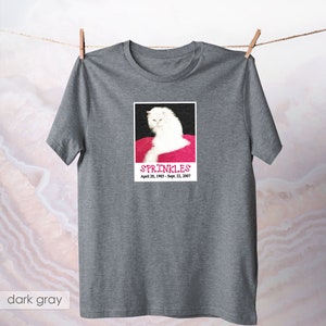 May include: A dark gray t-shirt with a white cat on a pink background. The cat is looking at the camera and the text "SPRINKLES" is printed below the image. The dates "April 20, 1985 - Sept. 22, 2007" are printed below the text.