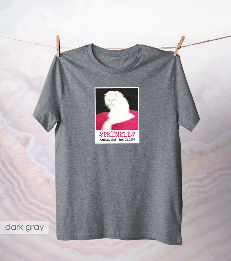 May include: A dark gray t-shirt with a white cat on a pink background. The cat is looking at the camera and the text "SPRINKLES" is printed below the image. The dates "April 20, 1985 - Sept. 22, 2007" are printed below the text.