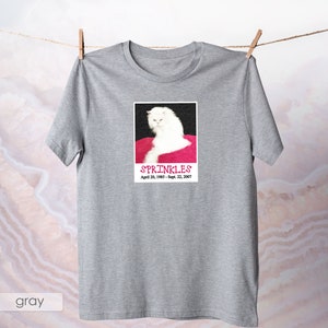 May include: Gray t-shirt with a white cat on a pink background. The cat is looking at the camera with a serious expression. The text "SPRINKLES" is printed below the cat, along with the dates "April 20, 1985 - Sept. 22, 2007".
