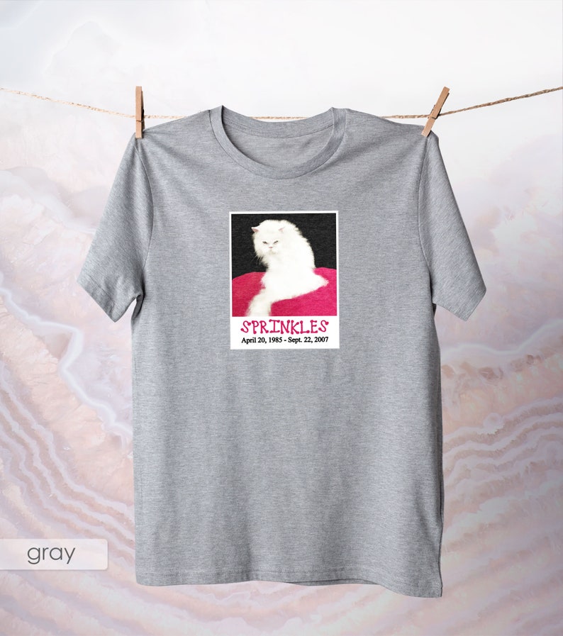 May include: Gray t-shirt with a white cat on a pink background. The cat is looking at the camera with a serious expression. The text "SPRINKLES" is printed below the cat, along with the dates "April 20, 1985 - Sept. 22, 2007".