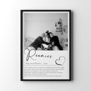 May include: A black and white framed wall art print featuring a photograph of three people sitting on a bed. Below the photo, the text reads Roomies, Meg, Jen & Harriet, noun, followed by a definition of friendship and a heart icon.