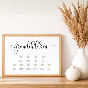 Op de afbeelding: Een rechthoekige houten lijst met een witte print waarop het woord grandchildren in zwarte cursieve letters staat. Onder de tekst staan tien beige hartjes in twee rijen, elk met een naam erbij: Jessie, Mikey, Lily, Ellie, Noah, Tommy, Hunter, Joseph, Melly en Theo.