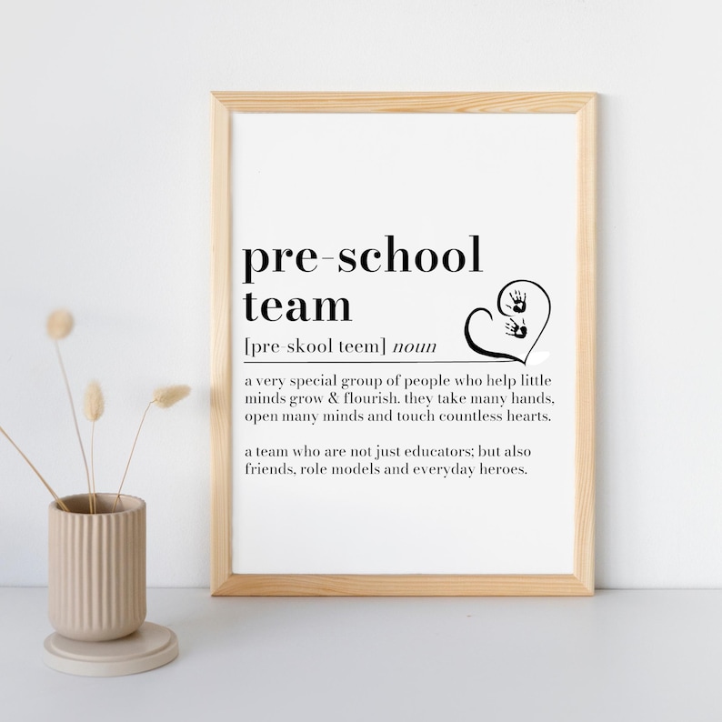 K&ouml;nnte beinhalten: Gerahmter Druck mit den Worten "pre-school team" in schwarzer Schrift. Die Definition des Begriffs steht unter dem Titel. Eine Herzform mit Handabdr&uuml;cken befindet sich rechts vom Text. Eine beige Vase mit Trockenblumen befindet sich im Vordergrund.