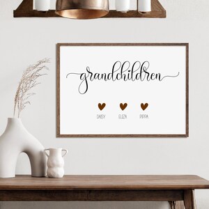 Op de afbeelding: Een rechthoekige wandkunstprint in een donkerhouten lijst met het woord grandchildren in zwarte cursieve letters. Onder de tekst staan drie bruine hartjes, elk geplaatst boven de namen Daisy, Eliza en Pippa in een eenvoudig zwart lettertype.