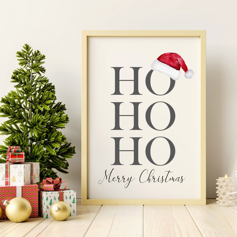 Puede incluir: Una impresi&oacute;n enmarcada con fondo blanco y texto gris que dice "HO HO HO Feliz Navidad". Un gorro de Pap&aacute; Noel rojo con ribete blanco se coloca sobre el texto.