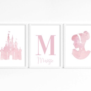 May include: Three pink watercolor prints in white frames. The left print features a pink castle silhouette. The middle print features a large pink letter M with the name Maisie below. The right print features a pink silhouette of a woman with a bun in her hair.