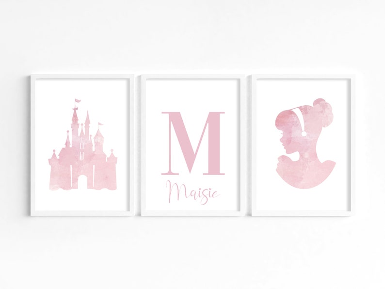 May include: Three pink watercolor prints in white frames. The left print features a pink castle silhouette. The middle print features a large pink letter M with the name Maisie below. The right print features a pink silhouette of a woman with a bun in her hair.