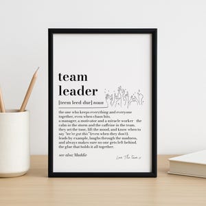 Puede incluir: Un marco negro con la frase "team leader" en negrita. La definición del rol de un líder de equipo está impresa debajo. El marco está sobre una superficie de madera.