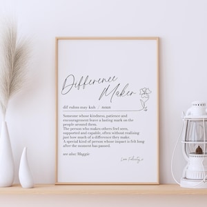 May include: A rectangular wall art print in a light wood frame featuring the word Difference Maker in black script, followed by its phonetic pronunciation, noun classification, and a definition of the term, ending with see also: Maggie and Love Felicity.