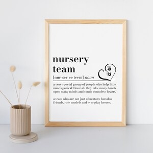 K&ouml;nnte beinhalten: Ein gerahmter Druck mit den Worten "nursery team" in schwarzer, fetter Schrift. Die Definition von "nursery team" steht unter dem Titel. Ein Herz mit zwei Handabdr&uuml;cken befindet sich auf der rechten Seite des Drucks. Eine beige Vase mit Trockenblumen ist im Vordergrund.
