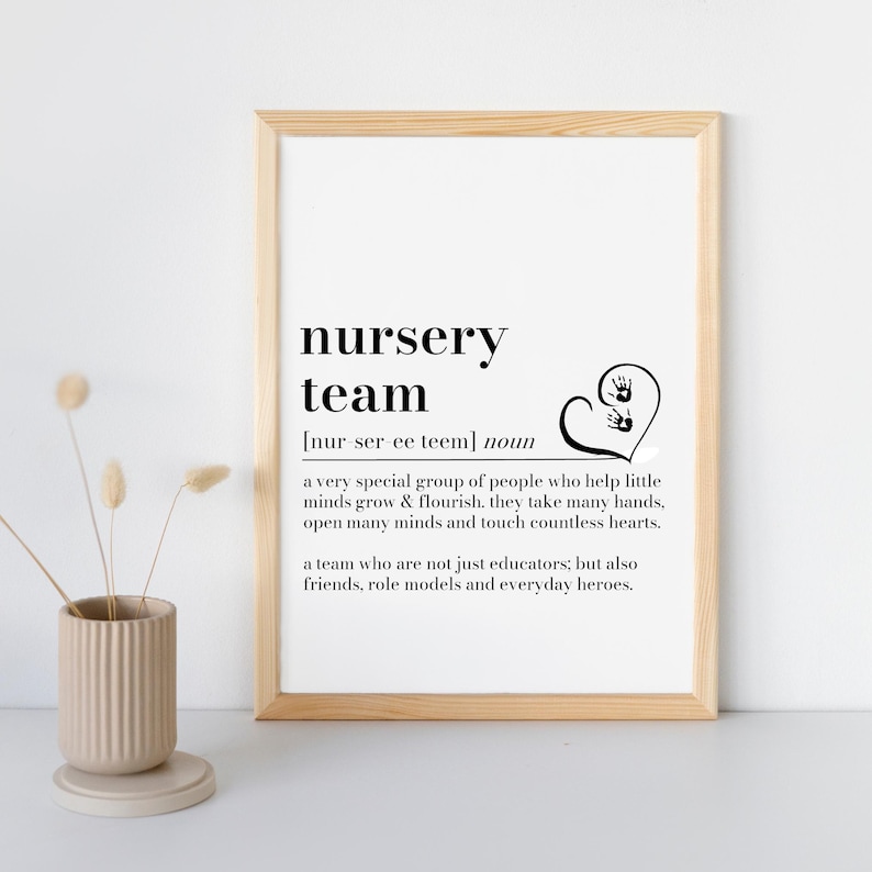 K&ouml;nnte beinhalten: Ein gerahmter Druck mit den Worten "nursery team" in schwarzer, fetter Schrift. Die Definition von "nursery team" steht unter dem Titel. Ein Herz mit zwei Handabdr&uuml;cken befindet sich auf der rechten Seite des Drucks. Eine beige Vase mit Trockenblumen ist im Vordergrund.