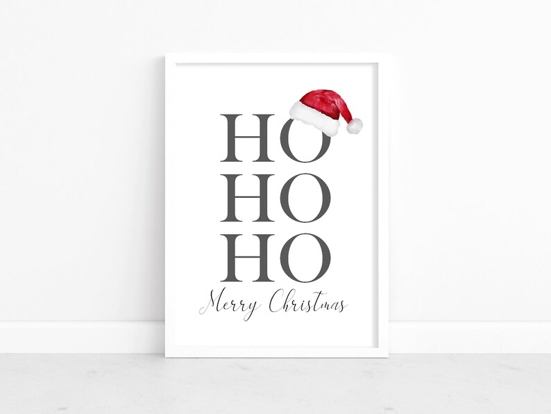 Puede incluir: Una impresi&oacute;n enmarcada en blanco con las palabras "HO HO HO Feliz Navidad" en texto gris. Un gorro de Pap&aacute; Noel rojo con ribete blanco est&aacute; en la parte superior de la impresi&oacute;n.