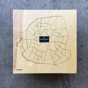 May include: A wooden jigsaw puzzle of the city of Charlotte, North Carolina. The puzzle is made by Puzzle Places and is on a light wood background.