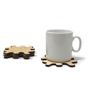 Puede incluir: Una taza de cerámica blanca se encuentra sobre un posavasos de madera con forma de pieza de rompecabezas. El posavasos está hecho de madera marrón claro y tiene un borde marrón oscuro.