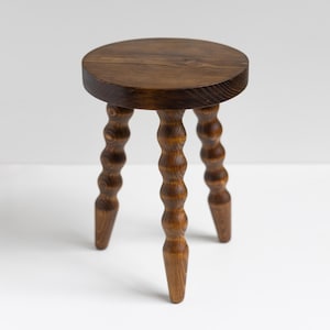 May include: A small, round wooden stool with a dark brown finish. The stool has a circular seat and three legs with a unique, turned bobbin design. The stool is approximately 30 cm tall.
