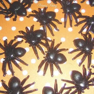 May include: A collection of black plastic spiders scattered on an orange background with white polka dots. The spiders have a rounded body and multiple legs, perfect for Halloween decorations.