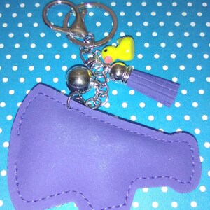 Op de afbeelding: Een paarse sleutelhanger in de vorm van een megafoon met een zilveren sleutelring en ketting. Het heeft een gele eendenbedel, een zilveren bal en een paarse kwast. De sleutelhanger staat op een blauwe achtergrond met witte stippen.