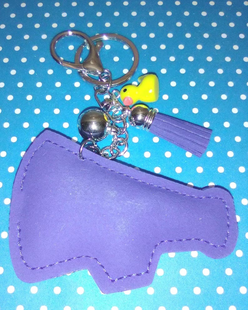 Op de afbeelding: Een paarse sleutelhanger in de vorm van een megafoon met een zilveren sleutelring en ketting. Het heeft een gele eendenbedel, een zilveren bal en een paarse kwast. De sleutelhanger staat op een blauwe achtergrond met witte stippen.
