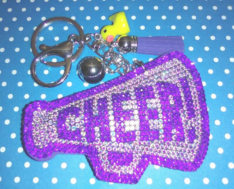Op de afbeelding: Een paarse cheerleader megafoon sleutelhanger met het woord "CHEER" uitgespeld in zilveren strass-steentjes. De sleutelhanger bevat een zilveren sluiting, een gele rubberen eend bedel, een paarse kwast en een zilveren bal bedel.