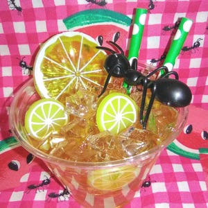 May include: A clear plastic cup filled with ice and a yellow-brown liquid, garnished with lime and lemon slices. Two green and white polka dot straws and a black ant figurine are in the cup. The background is a pink and white checkered pattern with watermelon slices and ant illustrations.