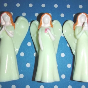 May include: Three small angel figurines. Each angel has a light green dress, white face and arms, and orange hair. The angels have large wings and are in different poses, with their hands clasped or holding a small pink object.