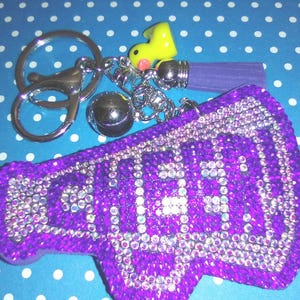 Op de afbeelding: Een paarse sleutelhanger in de vorm van een megafoon met het woord "CHEER" in heldere strass-steentjes. De sleutelhanger heeft een zilveren sleutelring, een kleine gele eendenbedel, een zilveren bal en een paarse kwast. De achtergrond is blauw met witte stippen.