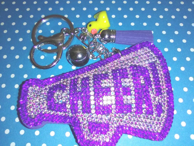 Op de afbeelding: Een paarse sleutelhanger in de vorm van een megafoon met het woord "CHEER" in heldere strass-steentjes. De sleutelhanger heeft een zilveren sleutelring, een kleine gele eendenbedel, een zilveren bal en een paarse kwast. De achtergrond is blauw met witte stippen.