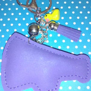 Op de afbeelding: Een paarse sleutelhanger in de vorm van een megafoon met een zilveren sleutelring en ketting. De sleutelhanger heeft een gele eendenbedel en een paarse kwast. De achtergrond is blauw met witte stippen.