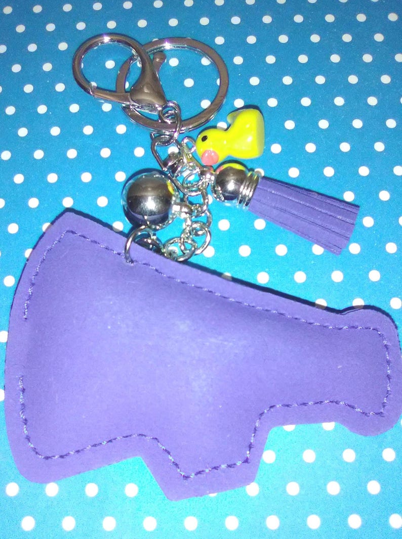 Op de afbeelding: Een paarse sleutelhanger in de vorm van een megafoon met een zilveren sleutelring en ketting. De sleutelhanger heeft een gele eendenbedel en een paarse kwast. De achtergrond is blauw met witte stippen.