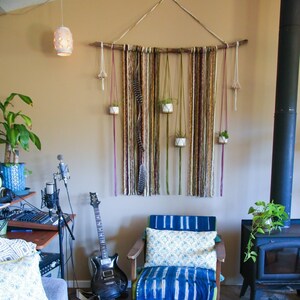 Pode incluir: Um interior de sala de estar com uma poltrona listrada azul e branca, uma mesa de centro de madeira, um violão e uma parede de macramé com plantas.