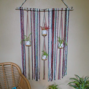 May include: A macrame wall hanging with four small white pots containing plants. The hanging is made with a variety of colourful yarns and is attached to a driftwood branch. A wicker chair with a blue and gold patterned cushion sits in front of the hanging. A small wooden table with a carved design sits next to the chair. A lit candle and a purple crystal are on the table. A potted plant with green leaves and a blue and white patterned pot sits on the floor.