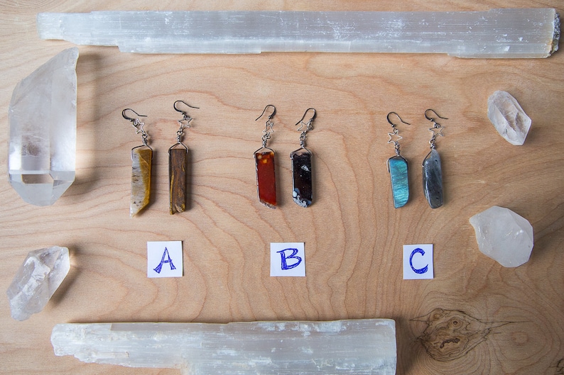 May include: Three pairs of dangle earrings with different coloured stone pendants. The earrings are labelled A, B, and C. The stones are tiger's eye, red jasper, and labradorite.
