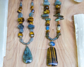Puede incluir: Dos collares con cordones de cuero marrón y cuentas de piedras preciosas. El collar de la izquierda tiene un colgante de labradorita azul en forma de lágrima. El collar de la derecha tiene un colgante de ojo de tigre rectangular. Ambos collares están etiquetados con las letras A y B.