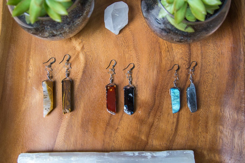 May include: Six pairs of dangle earrings with star charms. Each pair features a different semi-precious stone, including tiger's eye, agate, red jasper, black obsidian, labradorite, and grey agate.