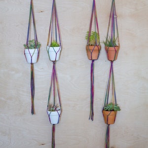 May include: Four macrame plant hangers with different colored strings, each holding a small potted plant. The hangers are hung on a light wood wall.