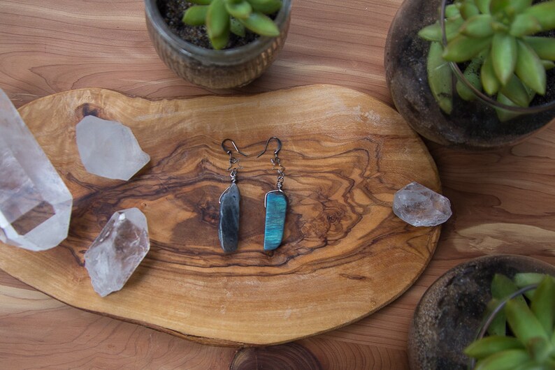 May include: A pair of dangle earrings with a dark grey stone on one side and a blue-green stone on the other side. The earrings are displayed on a wooden surface with clear quartz crystals.